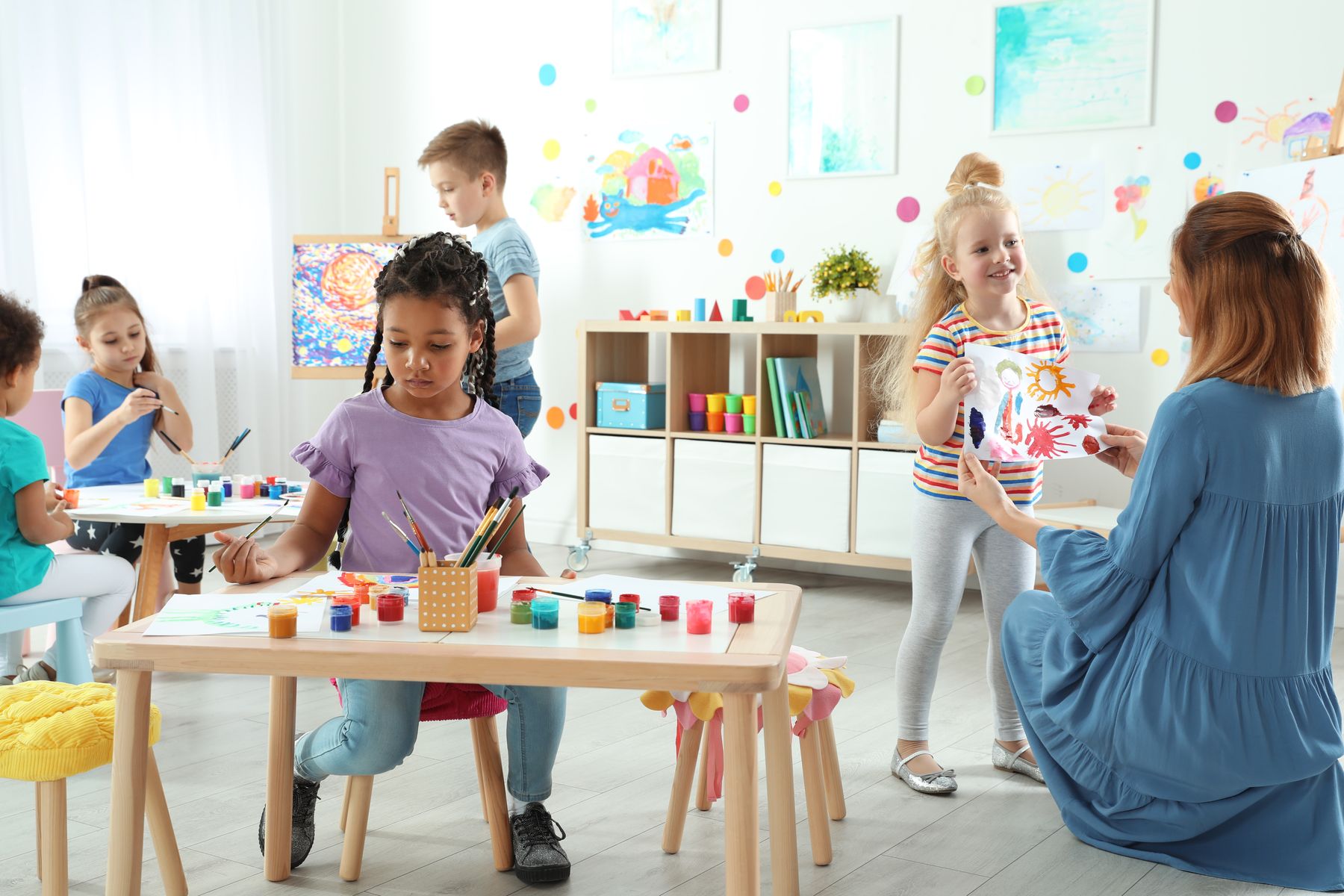 A child proudly shows her painting to an educator in a bright art room