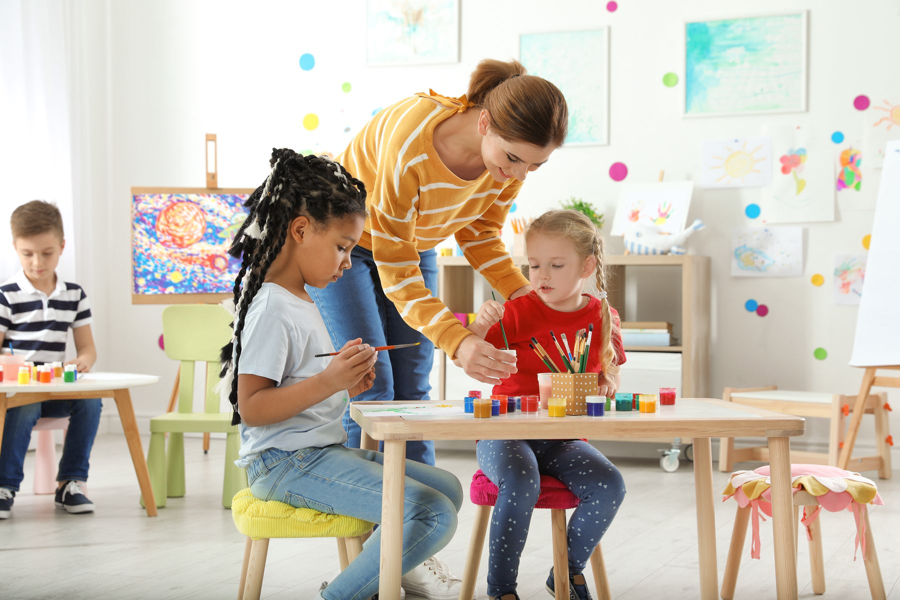 An educator leans in to help two young children painting at a classroom table