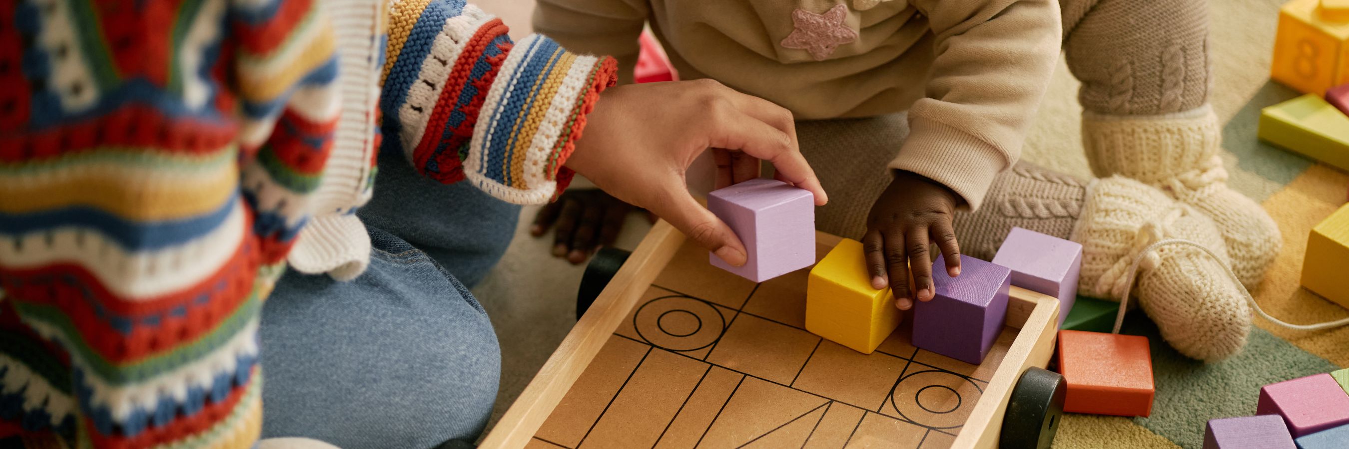 An infant and an adult place wooden blocks into a wooden cart together